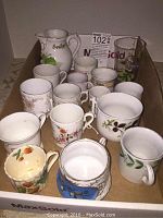 Box containing over 10 vintage cups and a ceramic pitcher, showing general condition and variety of items.