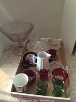 Full lot of various pressed glassware including trifle bowl, lidded candy dish, colored souvenir glasses, and white ceramic pitcher all arranged in box.