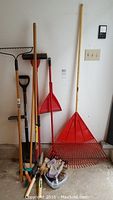 Collection of garden tools leaned against a wall including multiple rakes, a metal shovel, a broom, and gardening gloves on the floor.