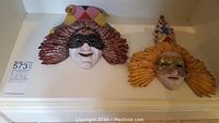 Two ceramic masquerade masks on display, one with purple/red hair and jester hat, one with yellow hair and colorful pointed hat.
