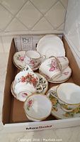 Photo showing multiple Royal Albert cups and saucers in a box, featuring different floral patterns with gold trim.