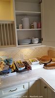 Kitchen cabinet with white plates, bowls, mugs, cutting boards, and plastic trays with kitchenware on countertop