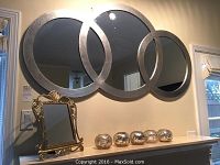 Wide shot showing the large wall mirror with three intersecting circular panels in metallic silver frame above a surface holding an ornate gold-tone freestanding mirror and five small silver candle holders.