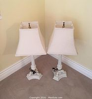 Pair of white painted metal column table lamps with white rectangular fabric shades, placed on carpeted floor against corner walls.
