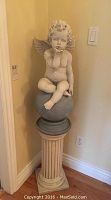 Full view of the cherub sculpture sitting atop the pedestal in a corner of a room with hardwood floors and beige walls.