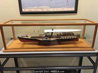 Side view of the handcrafted wooden paddle wheeler ferry boat model in oak and glass display case on a table against wall.