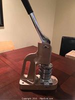 Side view of beige metal button maker press with chrome handle lever and circular dies attached, showing overall device design.