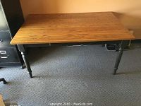 Front view of solid oak dining table with black base and golden oak top placed on carpeted floor.