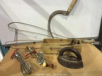 Full view of six antique items including flat iron, two mixers, wooden cheese box, wooden rug beater, and metal sickle laid out on a cardboard surface