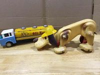 Two vintage toys displayed on wooden surface: yellow and blue tin oil tanker with some rust, missing one rubber wheel; plastic dog figurine with yellow hat and missing tail.
