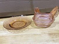 Two rose pink glass dishes shown side by side: a chicken-shaped covered dish with lid on the right and a round decorative plate on the left, both on a wooden surface.