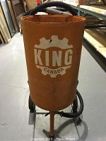 Front view of orange cylindrical sandblast pot with King Canada logo, showing hose and wheel base.