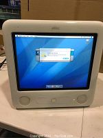 Front view of the vintage Apple eMac computer powered on, screen showing OS X interface with a popup window. White plastic casing, built-in stereo speakers below screen.