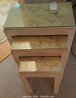 Front view of the 3 nesting tables stacked, showing carved lilies under glass top on pickled wood tables.