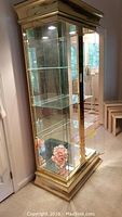Full view of the Design Institute of America curio cabinet in brass finish with glass shelves and top lighting, showing clear glass door and reflective mirrored back panel.