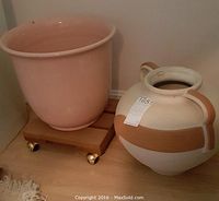 Photo showing the 12-inch pink plant pot on a rolling wooden stand with brass casters next to a 12-inch terra cotta urn with white decorative bands and handles.