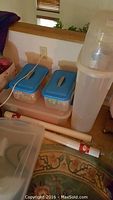 Overall lot showing tall plastic wrapping paper storage tube, several plastic containers including two with blue lids, and rolls of wrapping paper on floor.