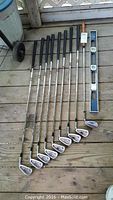 Full set of nine Square Two Powecircle XL golf irons along with Wilson SW 55 Tour TC2 wedge, a wooden handled metal level, and a single ski pole visible on a wooden surface