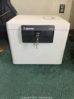 Front view of the white Sentry portable safe placed on green carpet, showing the black locking panel and keys inserted.