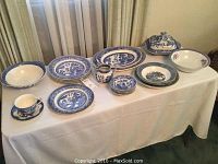 Full display of the partial china set arranged on a table showing plates, bowls, cup and saucer, and serving dish with blue and white scenic patterns.
