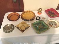 Top-down view of eight vintage glass ashtrays in various shapes and colors along with two lighters on a white tablecloth.