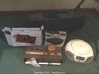 Wide photo showing all items in the lot: wood-grain clock radio with microphone, white portable CD player, black GPC clock radio, digital clock radio with red LED display in box