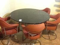 Full view of round glass top table with five retro style chairs in red-orange upholstery and metal frames.
