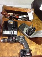 Photo showing cordless drill with extra battery, charger, metal case containing drill bits, and wooden tray with various hand tools