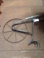 Metal hand cultivator with circular spoked wheel and three curved tines lying on floor