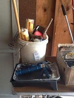 White bucket filled with paint rollers, roller handles, masking tape rolls and paint brushes, with a paint tray and blue caulking gun in front