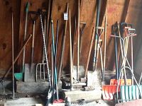 Full view of garden tools including shovels, pitchfork, axes, and sledge hammer leaning against a wood wall.