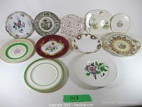Eleven assorted vintage display plates laid out on a surface showing various sizes and floral/special designs with minor wear.