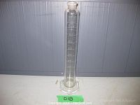 Full view of the tall clear glass measuring cylinder with volume markings and hexagonal base on a gray background.