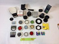 All camera and lens accessories spread on white surface, including Canon EF 50mm f/1.8 II lens, flash diffuser, multiple filters in plastic cases, tripod clamp, and memory card.