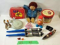 Wide shot showing the ALF lunch bag, Cabbage Patch doll, Mr. Potato Head, Star Trek USS Enterprise, small figurines, sunglasses, toy lightsabers, and pink bead bracelet.