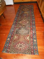 Full length view of the old area rug on hardwood flooring showing its pattern, colors, and worn fringes.