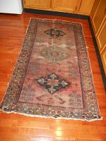Full view of rectangular woven rug on hardwood floor. Shows entire pattern, color, and wear including frayed edges.