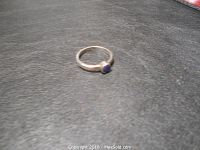 Ring marked 925 with a small round blue stone set on a simple silver band, shown from an overhead angle on a black textured surface.