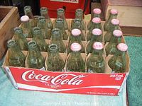 24 Coca-Cola Classic glass bottles in a cardboard case with some caps and visible wear.