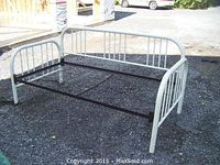 White painted metal daybed frame with rounded headboard and footboard with vertical bars, black metal central support, shown on gravel surface.