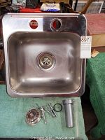 Top view of square stainless steel sink with two mounting holes and visible drain components placed beside it.