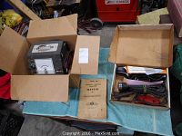 Top angled view of the Merc Tronic Model 98 analyzer in a cardboard box beside the Merc Tronic timing light set in its box with manuals laid out on a blue cloth on a table.