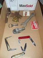 Overview of lot inside cardboard box showing knives, keys, forks, bell door knocker, bottle opener, metal scoop, wrench, and patch.