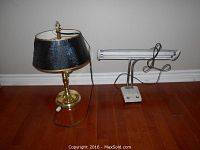 Both the brass table lamp with black shade and the fluorescent desk lamp side-by-side on wooden floor in front of a light gray wall.
