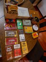 Photo showing collection of vintage tobacco pouches and tins on dark wood table including Players, Black Cat, Amphora, Hollandia, Sweet Virginia, Sail, and Flying Dutchman brands, all showing wear consistent with age.