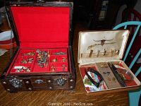 Open wood jewelry box showing multiple compartments filled with men's jewelry including cuff links and tie clips, next to a smaller beige case with additional jewelry and watches.