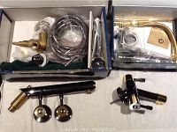 Two gunmetal grey faucets with gold plated accents in open boxes including hoses and installation parts.