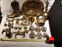 Wide shot showing the majority of the brass items laid out on white fabric including cowboy boots, large bowl, candelabra, ewer, multiple animal figurines, and plaques.