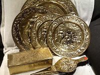 Photo showing four brass plates with embossed decorative scenes and floral borders along with a long rectangular matchbox with embossed design.