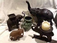 Five elephant figurines of different materials arranged on a light fabric background. The metal elephant is the largest and has a candle placed on its back.